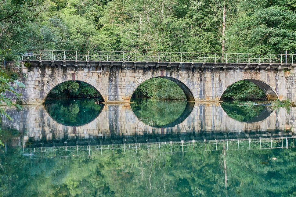 The image shows the photograph of a bridge with three arcades crossing a river. The bridge is reflected in the still water of the river.