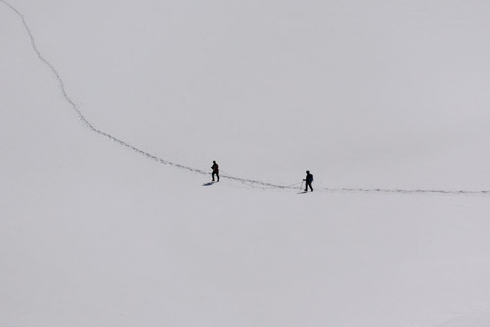 The image shows the photography of two hikers in a large field of snow somewhere in the Swiss Alps.