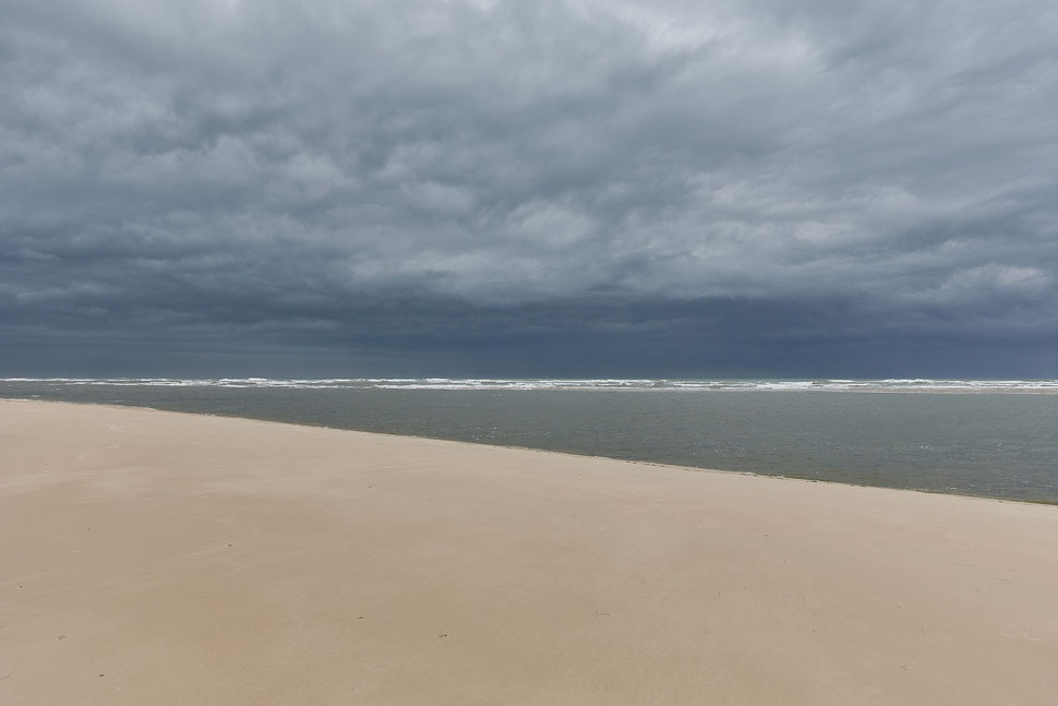 The image shows a photograph of a sandy beach on a stormy day. The water and clouds are rendered in various shades of gray. The location is an island close to Olhao.