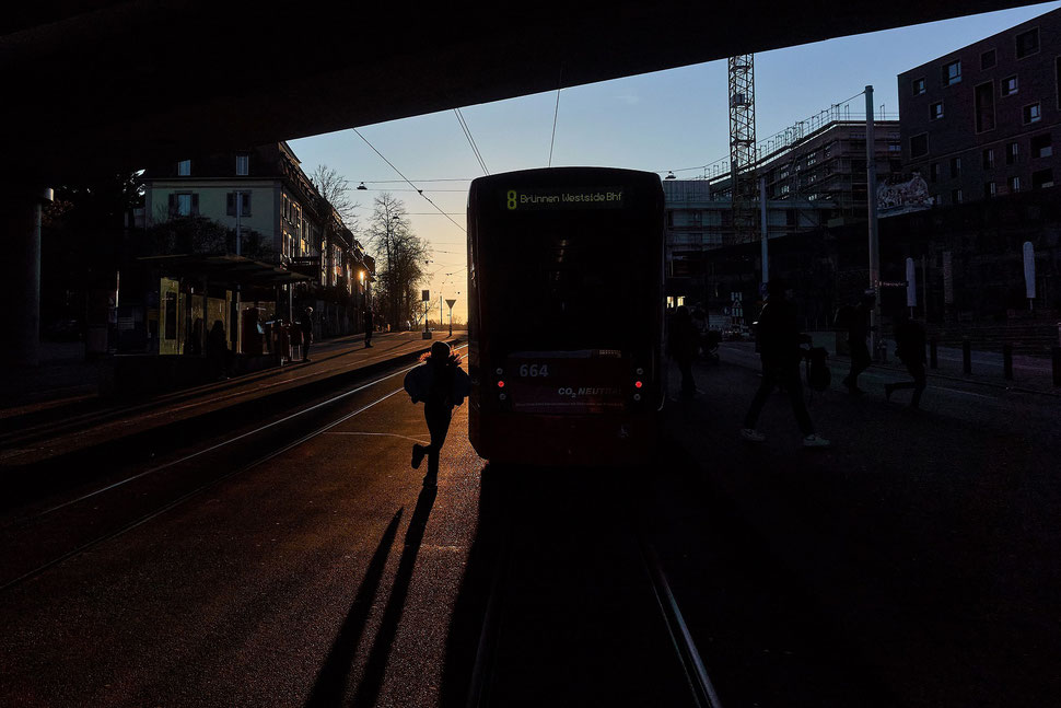 The image shows a woman running to catch tram no. 8 at Europaplatz in Bern while the sun is setting.