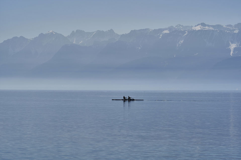 The image shows the photograph of boat with two people rowing in Lake Geneva against the backdrop the Swiss alps on a hazy morning.