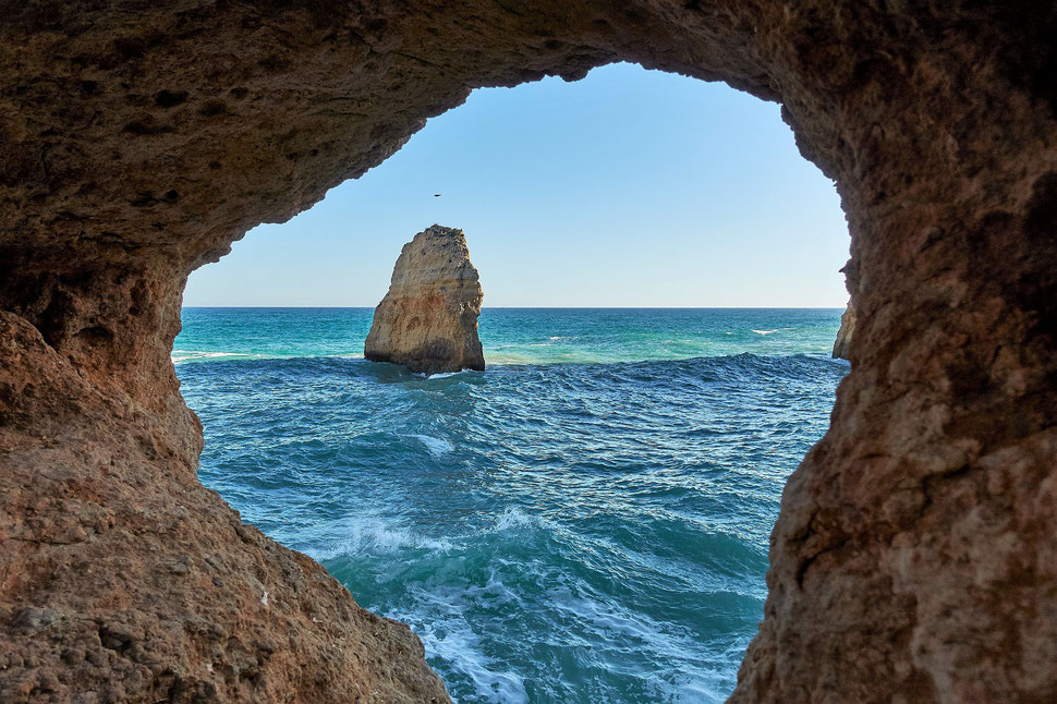 The image shows a photograph of a cliff hole through which you can see the sea and a protruding rock.