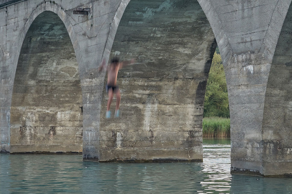 The image shows the photograph of a boy jumping from a bridge into the water of the Wohlensee (Lake Wohlen).