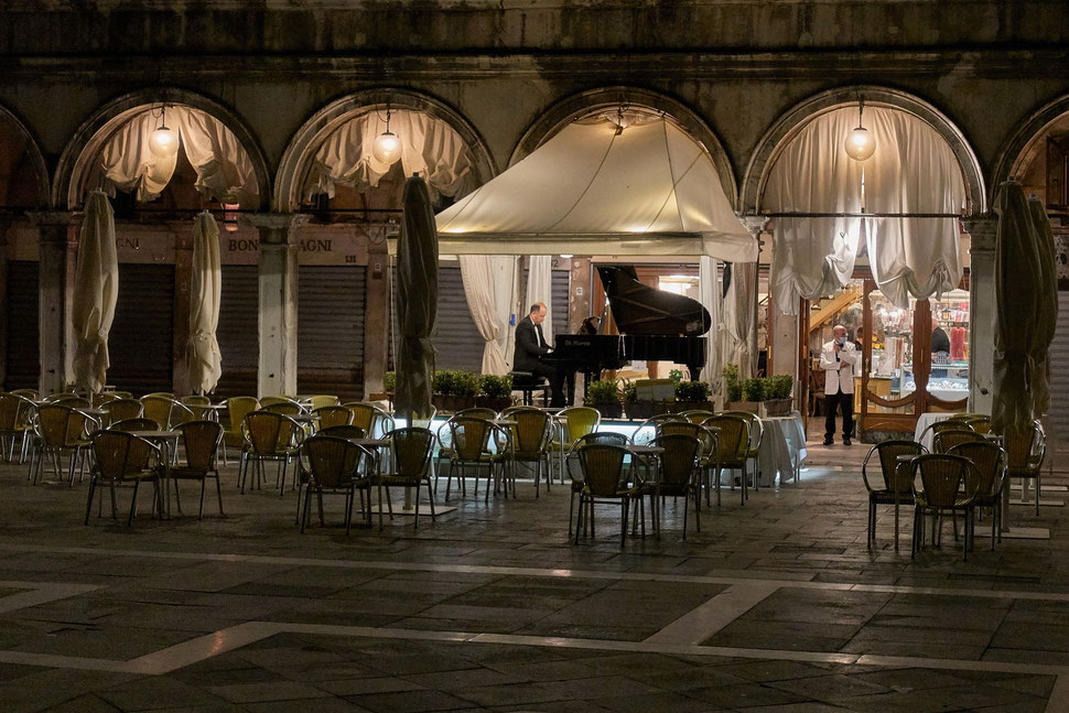 The image shows the nocturnal photograph of a waiting waiter and a piano player in an empty cafe in Venice.