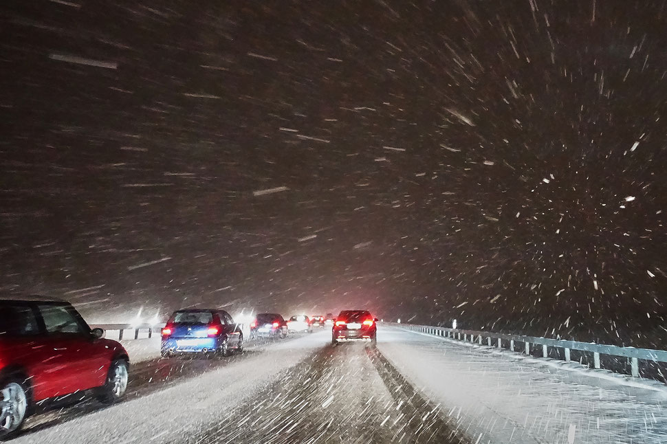 The image shows a nocturnal photograph of cars from behind on a snow covered freeway while it is heavily snowing. 