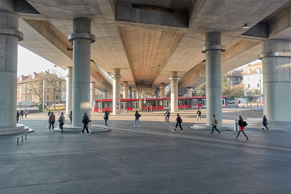 The image shows the photograph of Europaplatz Bern with some people crossing the square, a stopped red tram and a huge concrete overpass. 