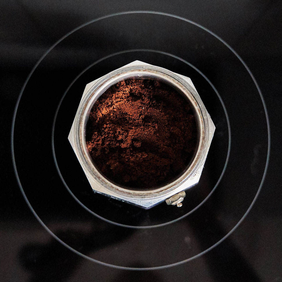 The image shows the photograph of an open Italian coffee maker with sunlight on ground coffee, which is placed on a cooking plate. 