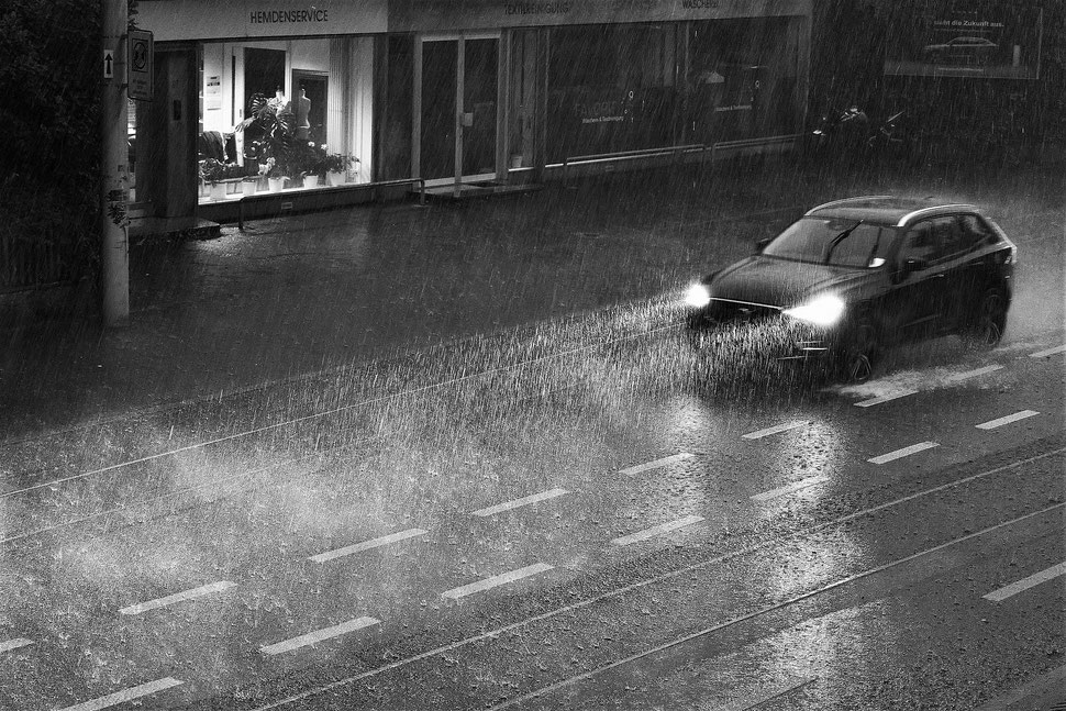 The image shows a nocturnal photograph of a car driving through a heavy rainstorm in front of a storefront and a billboard.