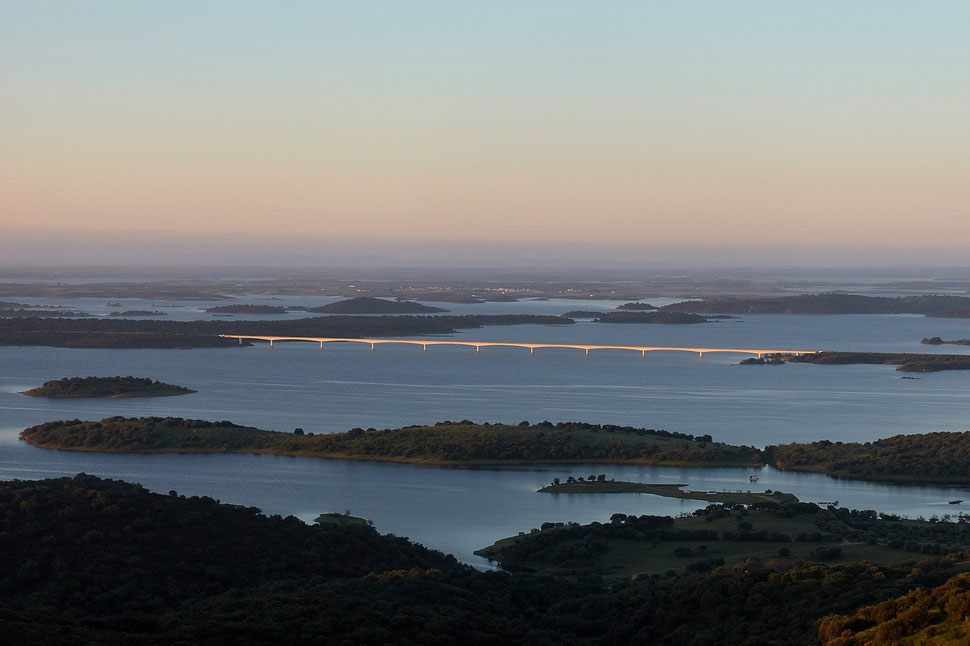 The image shows the photograph of a landscape consisting of small islands and areas of water; a bridge - illuminated by the morning sun - can be seen in the middle of the photograph.