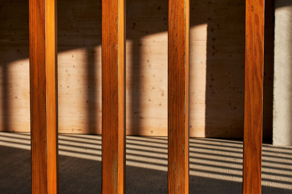 The image shows several wooden beams that are light by late afternoon sunshine and produce a pattern on the floor of a pavillion.