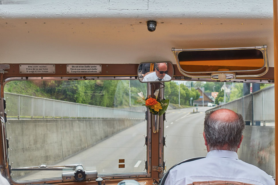 The image shows the photograph of the back of the head of a bus driver and the windshield with the road ahead. You can see the driver's face in the rear view mirror, which is placed in the center above the windshield. 