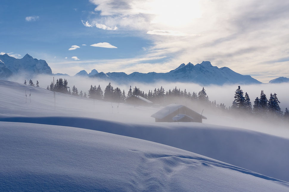 The image shows the photograph of an alpine pasture with a mountain hut in winter. A mountain range is visible in the background. The location is Balisalp (Hasliberg).