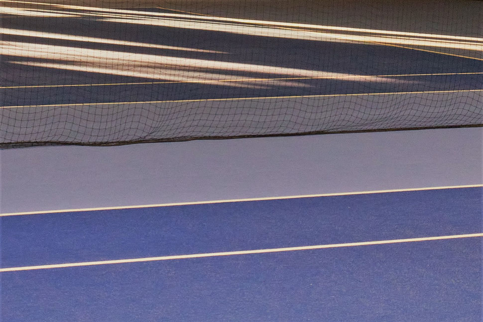 The image shows two indoor tennis courts that are seperated by a net. One of the courts is lit by early morning sunlight pouring into the hall.