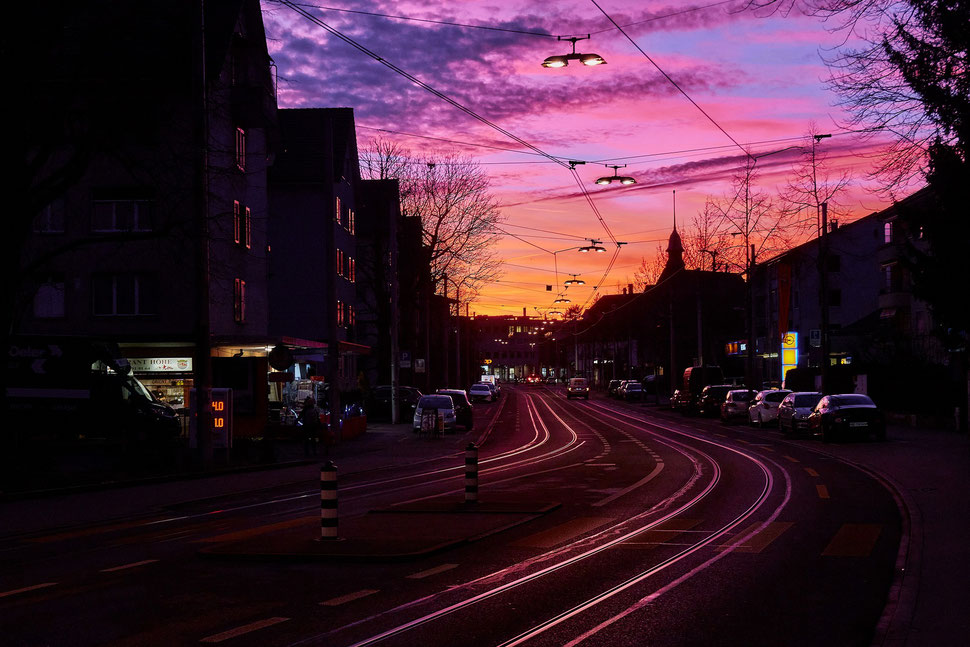 The image shows a photo of a sunset scene in the west of Bern - the tram tracks reflect the pink and orange light of the sky.