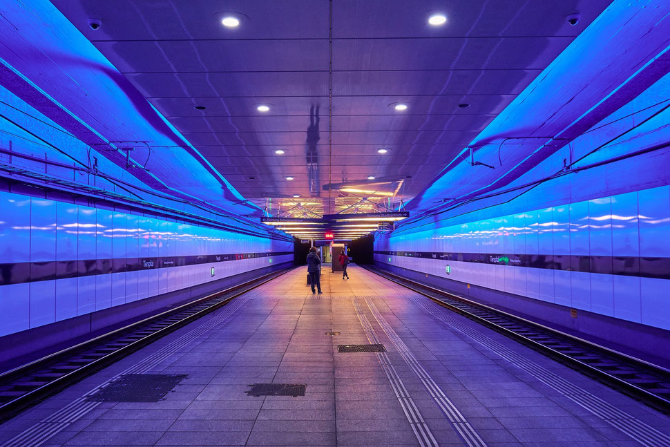 The image shows the photograph of two waiting people in a subway tram station in Zurich from a symmetrical perspective.   