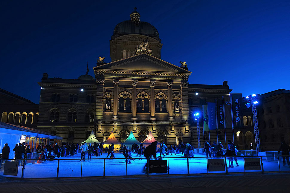 The photograph shows an atmospheric image of people skating on an ice skating rink in front of the federal palace in Bern.