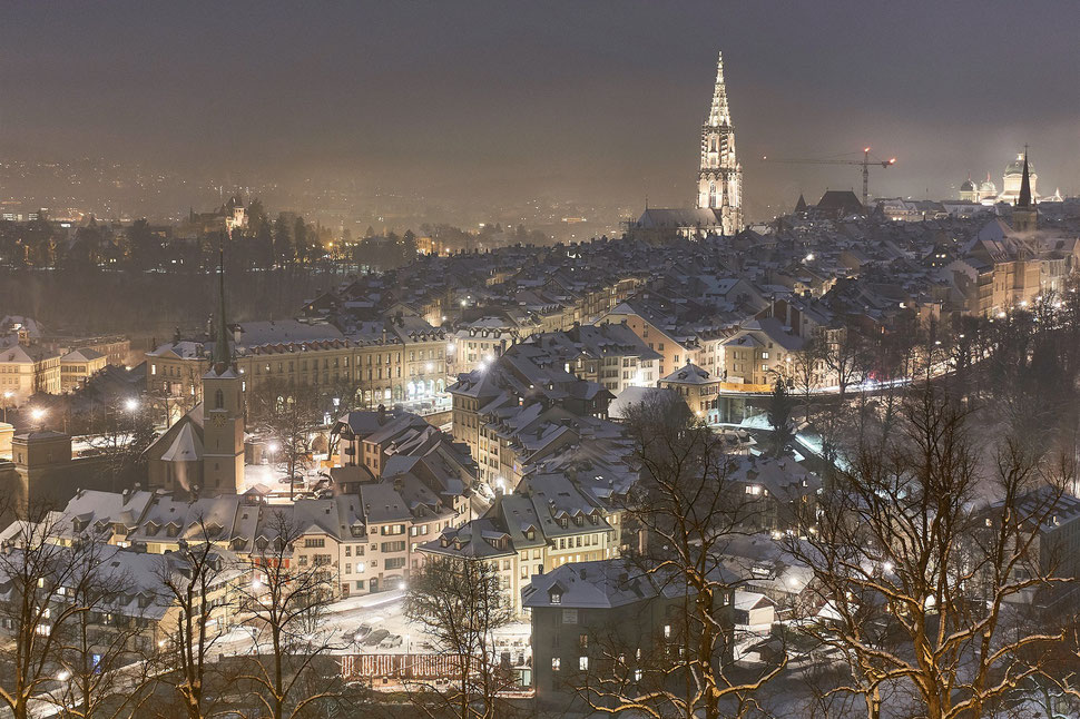 The image shows the nocturnal photograph of the city of Bern in Winter. The roofs are covered with snow and the streetlights light the city. 