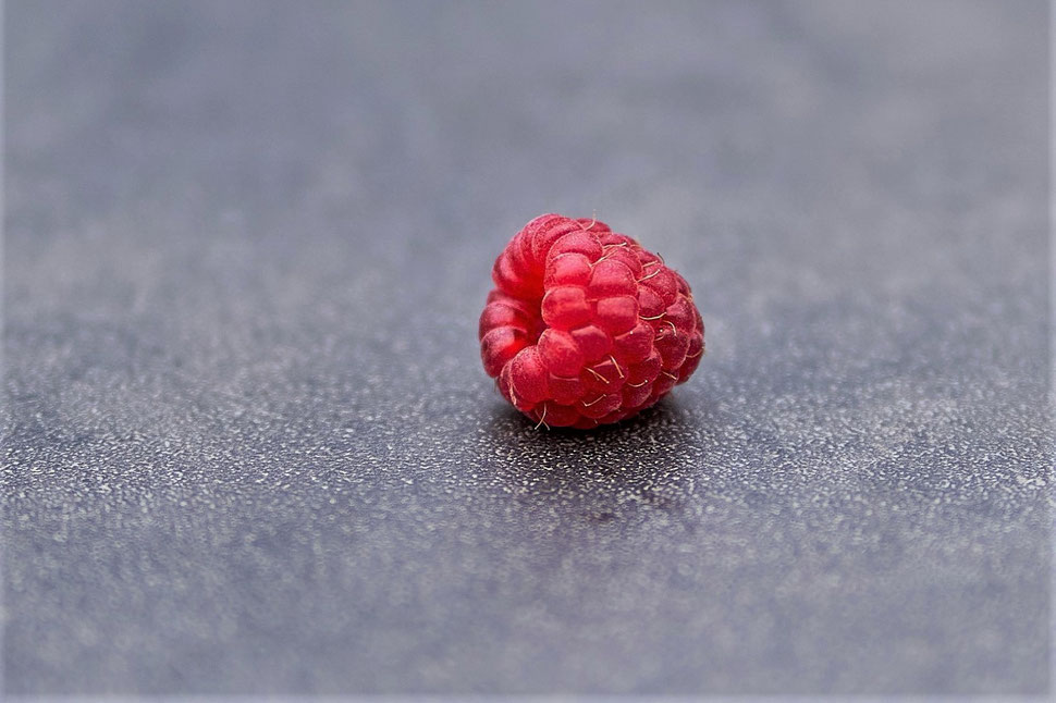 The image shows a macro photograph of a single raspberry on the surface of a table. 