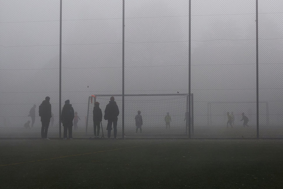 The image shows the photograph of kids playing soccer behind a high chainlink fence while their parents watch. It is a foggy Winter morning.