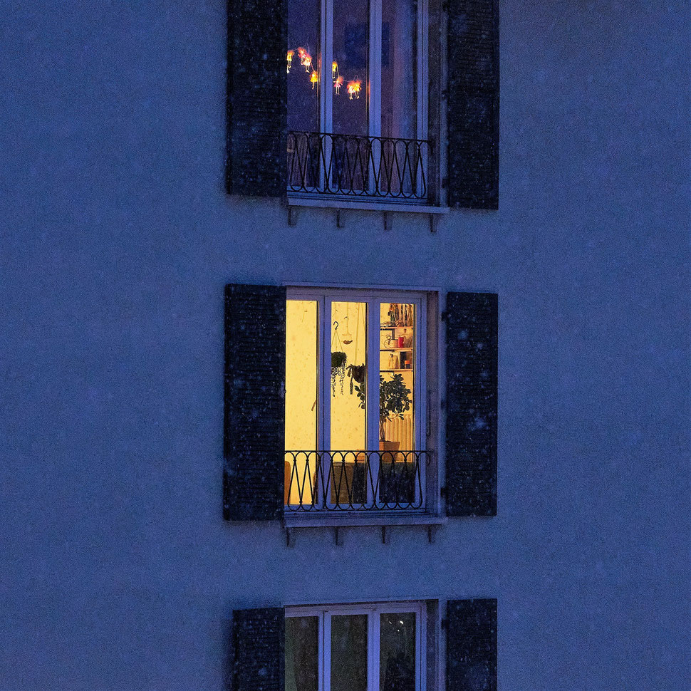 The image shows a photograph of a facade with several lit windows on a Winter night. It is lightly snowing.