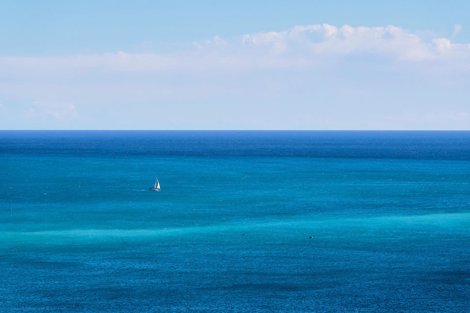 The image shows a photograph of a small sailing boat on the Mediterranean sea below a blue sky with white clouds. The sea water and the skyl boast different shades of blue.