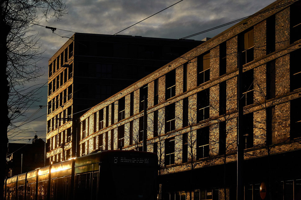The image shows the photograph of a large building an a passing tram - everything is illuminated by the warm light of the setting sun.