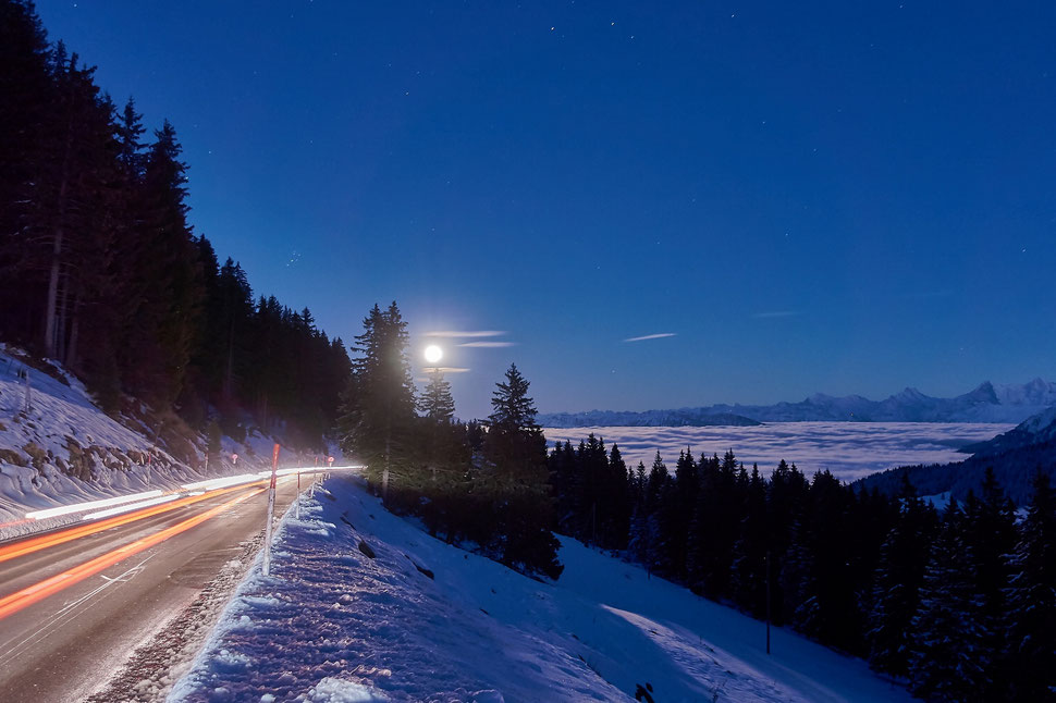 The image shows a photograph of a winterlandscape with mountains, fir trees and a full moon rising over the horizon. There's a road in the left part of the image with light tracks of cars that passed.