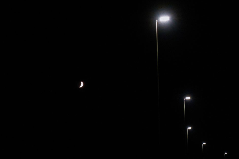 The image shows the black and white, minimalist photograph of an arrangement of five street lights and a small half moon against the backdrop of a black night. 