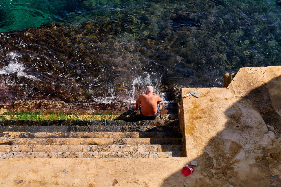 The image shows the photograph of an old man in swimming trunks sitting on the rocky steps of a sea bath in Santa Cesare Terme (Apulia). It is morning and everything is bathed in warm sunlight.