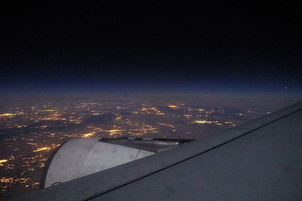 The image shows a night photograph through the window of an airplane; part of the wing, the lights of civilization and the dark sky are visible.