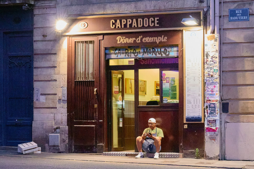 The image shows a photograph of a man sitting in front of his store in Bordeaux (France) taking a abreak and checking his smartphone.