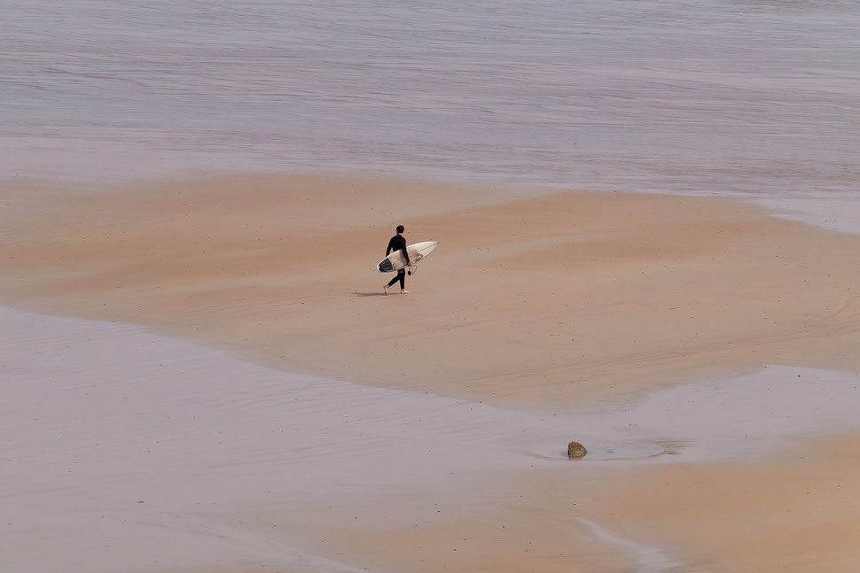 The image shows the minimalistic photography of a surfer carrying his surf board on a beach in Finistère (Brittany).