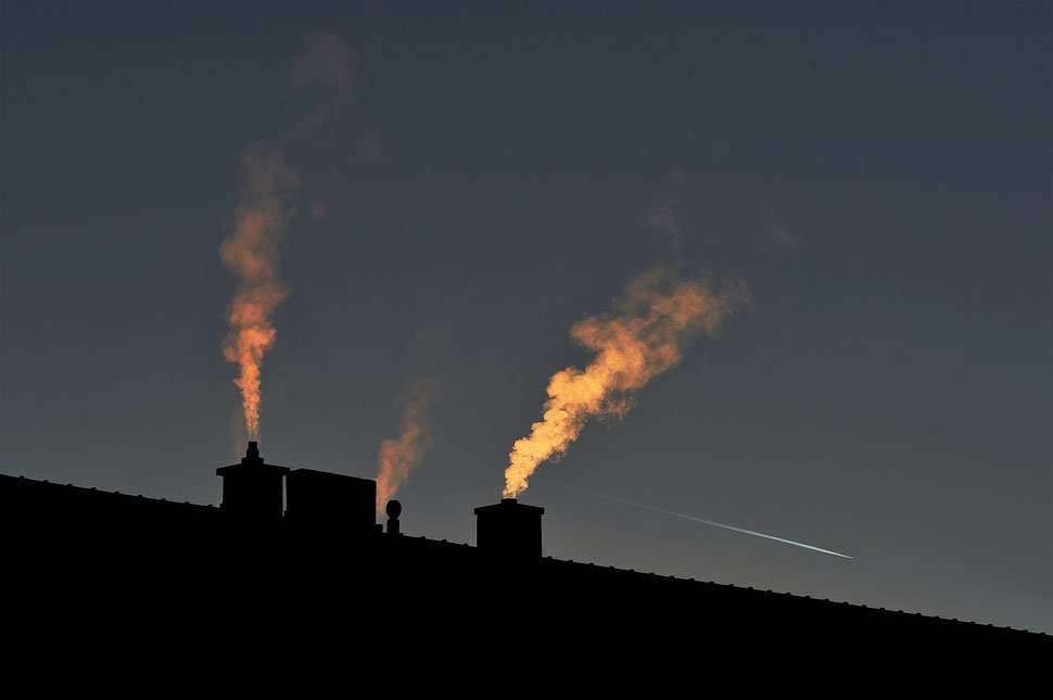 The image shows a photograph - taken against the light - of the outline of a roof with several smoking chimneys on a Winter morning. The sunlight colors the smoke in different shades of yellow and orange. 