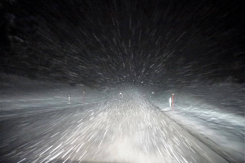 The image shows the night photograph of a snow coverd street lit by the headlights of a car and whirling snow flakes.