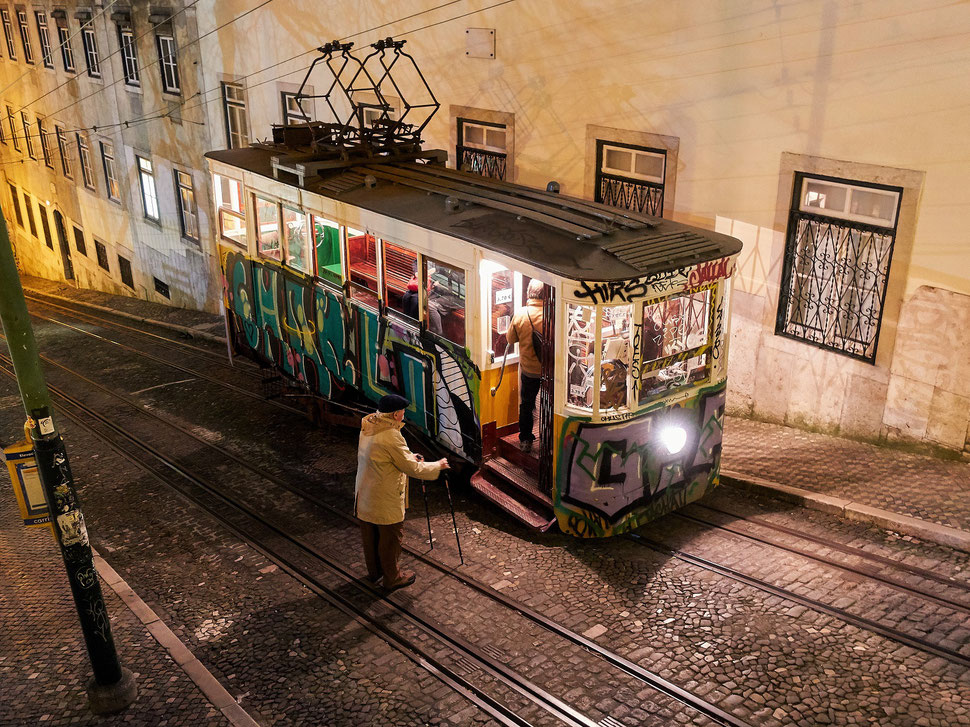The image shows a photograph of an old man getting ready to enter a Tram (Ascensor da Gloria) in Lisbon. It is night an the street is lit by street lights.