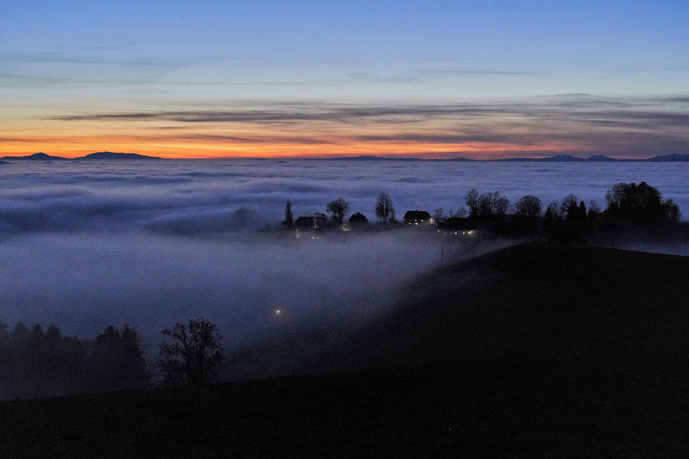 The image shows an evening photograph of a landscape with a sea of fog, some houses with lights, some trees and a colorful horizon.