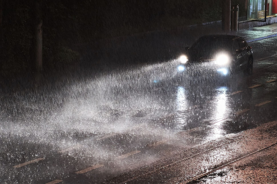 The image shows the photograph of a car driving through a heavy rainstorm in the night. The headlights light up the rain.