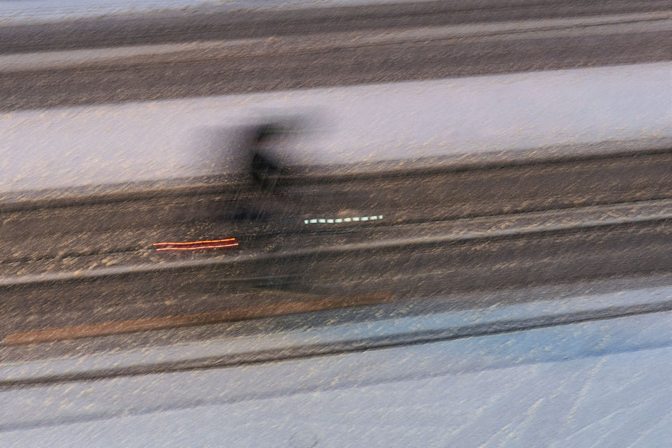 The image shows the photograph of the blurred shape of a cyclist on a snow covered street on a Winter evening.
