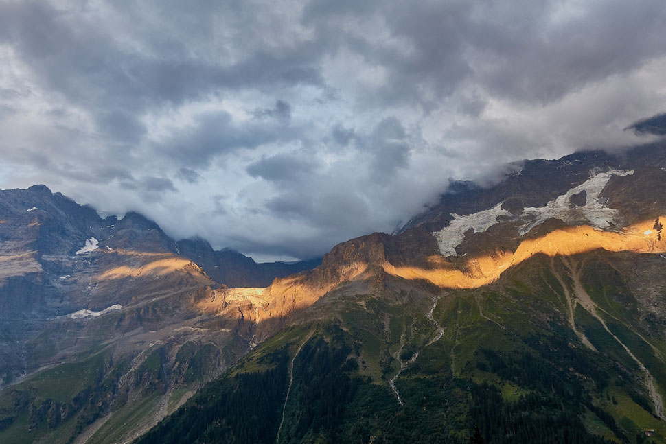 The image shows the photograph of mountains in the Bernese Oberland touched by a streak of evening sunlight under a clouded sky.