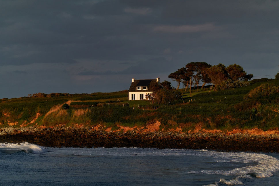 The image shows the photography of a white house on the coast of Brittany. The facade of the house and the white caps of the waves are illuminated by the setting sun.