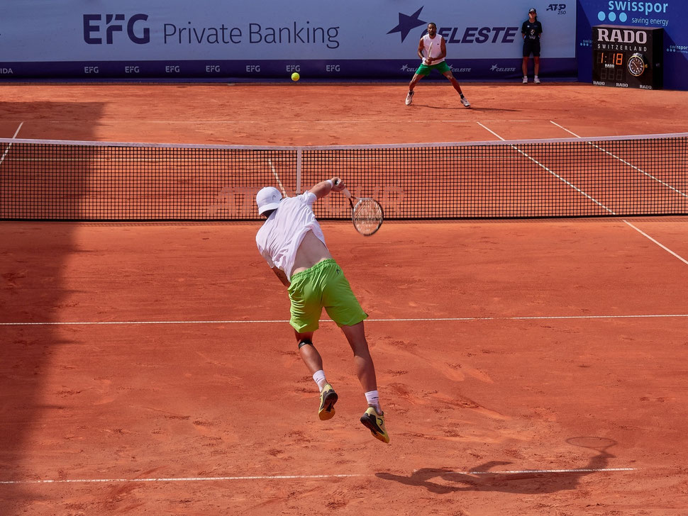 The image shows the photograph of the tennis player Jérôme Kym serving (from behind) at the Swiss Open 2025 in Gstaad. You can see his opponent - Calvin Hemery - getting ready to return.