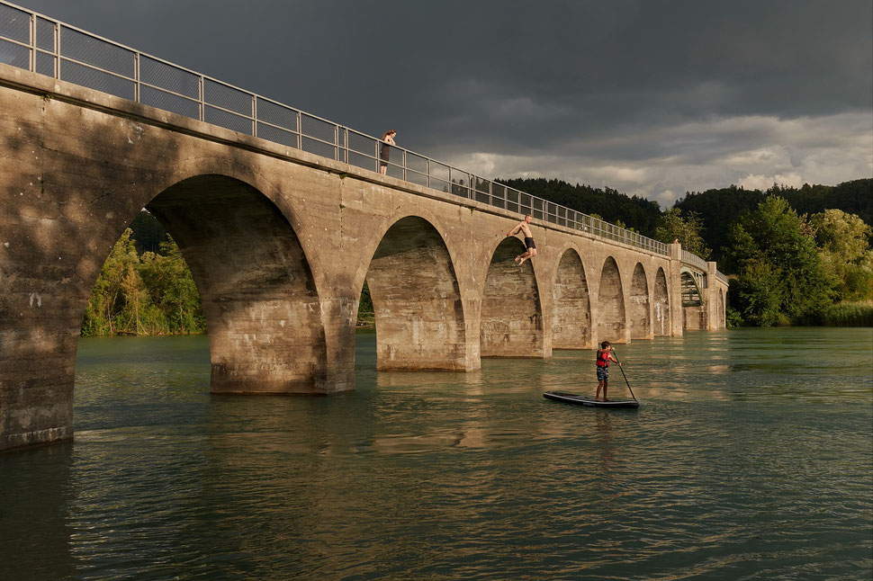 The image shows the photograph of a bridge, from which a young man - watched by a girl - is jumping into the water. It is a summer evening and a strom is brewing.