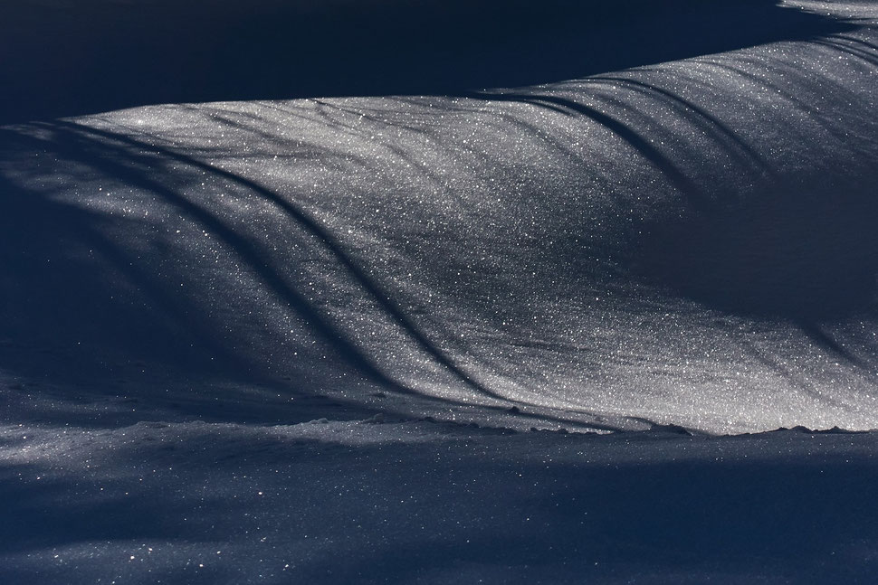 The image shows the photograph of a slope of snow that - in some places - sparkles in the sun and displays shadows in other places. 