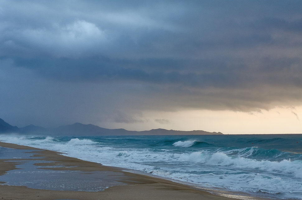 The image shows the photograph of a beach in Sardinia on a fall morning. Dark clouds and a range of mountains are visible in the background. 