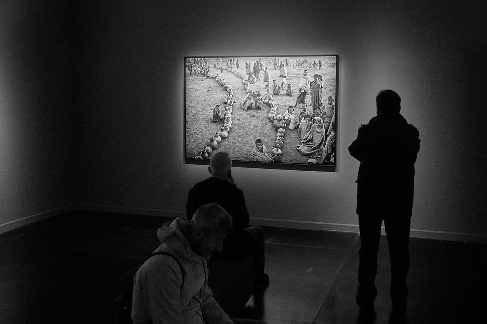 The image shows the photograph of sitting and standing people in a museum watching a print with african people sitting and standing.