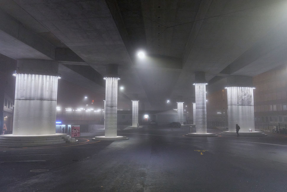 The image shows the night photograph of a man walking under a huge bridge with large lit pillars on Europaplatz, which is in the west of Bern.