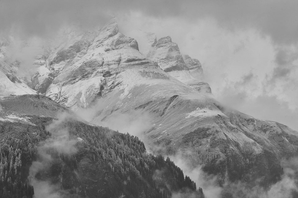 The image shows a black and white photograph of mountains that are partly hidden behind fog and lightly covered in snow.