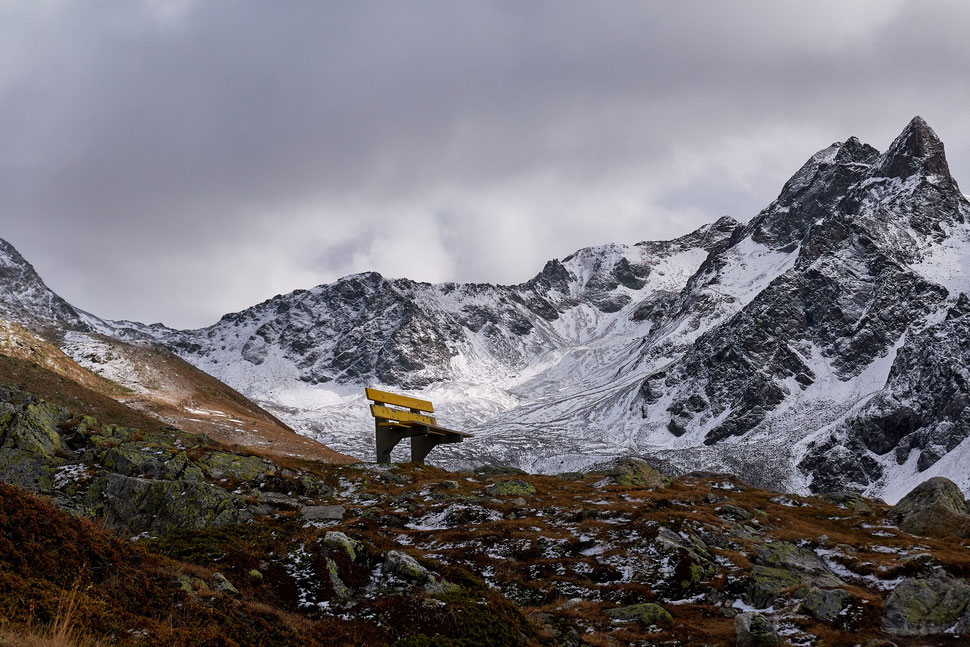 The image shows the photograph of a yellow bench in a mountainous setting under a grey sky. The location is Muottas Muragl (Engadin).