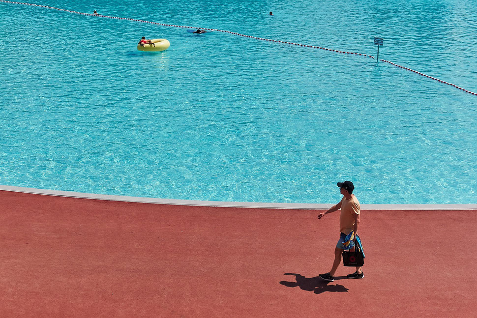 The image shows the colorful photograph of a large swimming pool with 4 people swimming in it. In the foreground a man in summer clothes passes the pool.