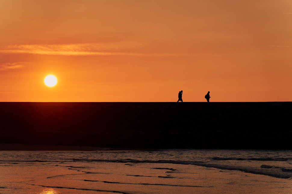 The image shows a photograph of two people walking on a pier while the sun is setting.